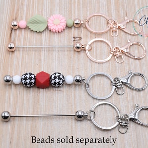 May include: Two sets of keychains with a silver and rose gold finish. Each set includes a ring, a swivel clasp, and a beaded bar. The beaded bar on the top set features pink, green, and white beads. The beaded bar on the bottom set features black and white patterned beads, a red bead, and a white bead. The text "Chews Me" is visible in the background. The text "Beads sold separately" is visible at the bottom of the image.
