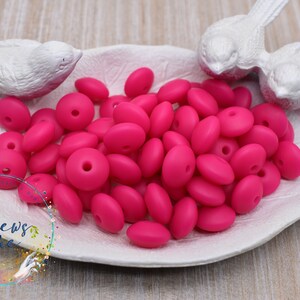 May include: A pile of bright pink silicone beads with a hole in the center. The beads are in a white dish with two white bird figurines on the edge.