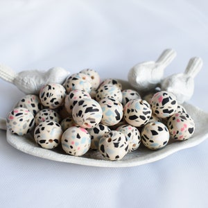 May include: A collection of round, white silicone beads with a black, pink, and blue abstract pattern. The beads are in a white ceramic dish.
