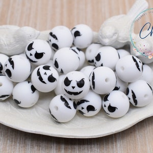 May include: A close-up of a pile of white silicone beads with black jack-o'-lantern faces. The beads are in a white dish with a bird-shaped handle. The text "Chews Me" is visible in the background.