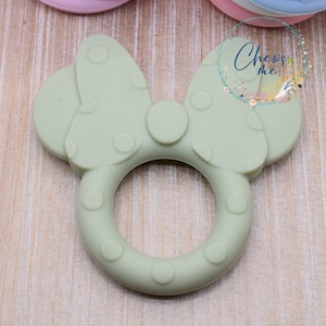 K&ouml;nnte beinhalten: Ein gr&uuml;ner Silikon-Bei&szlig;ring in Form einer Schleife mit wei&szlig;en Punkten. Der Bei&szlig;ring hat einen Ring in der Mitte zum einfachen Greifen.