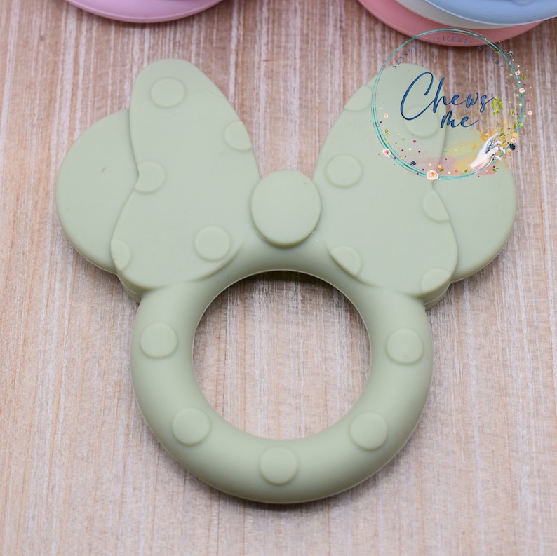 K&ouml;nnte beinhalten: Ein gr&uuml;ner Silikon-Bei&szlig;ring in Form einer Schleife mit wei&szlig;en Punkten. Der Bei&szlig;ring hat einen Ring in der Mitte zum einfachen Greifen.