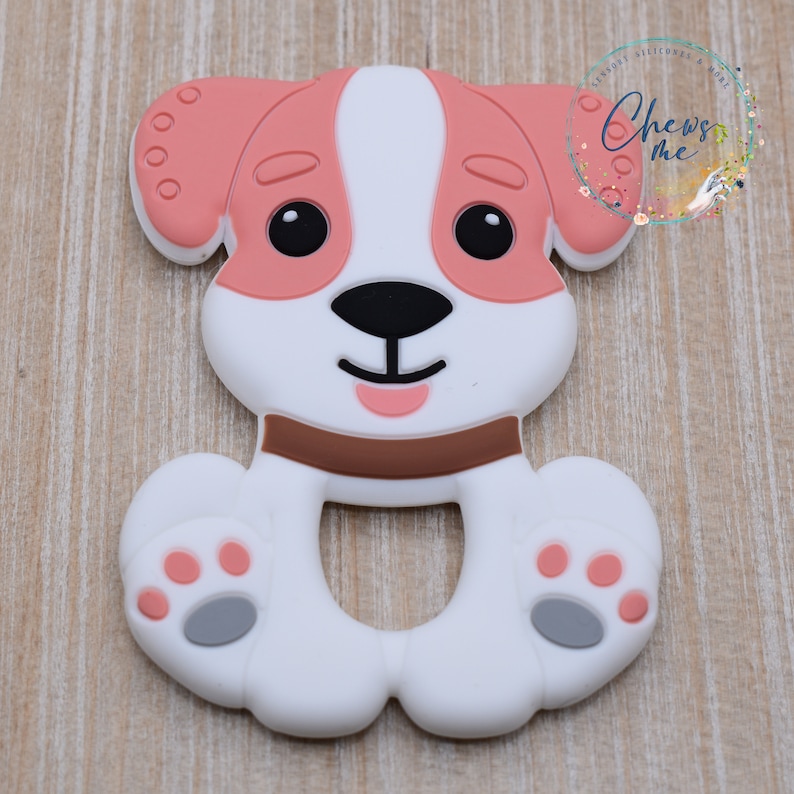 May include: A pink and white silicone teether shaped like a dog. The dog has a brown collar and gray paw prints.