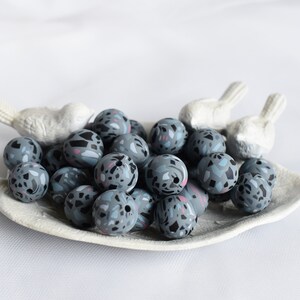 May include: A collection of gray and pink patterned beads in a white ceramic dish. The beads are round and have a textured surface.
