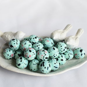 May include: A collection of light blue terrazzo beads with black and pink flecks, displayed on a white, leaf-shaped dish. Two white bird figurines are positioned behind the dish. The beads are spherical and appear to be handmade.