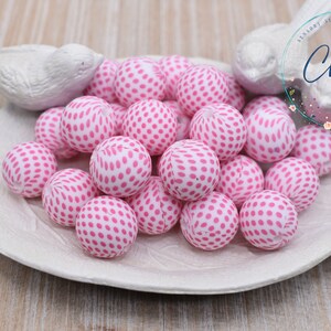 May include: A collection of pink and white dotted beads, displayed on a white, leaf-shaped dish. The beads are round and have a pattern of pink dots. The dish is flanked by two white bird figurines. The image includes the text "Chews me".