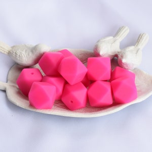 May include: A close-up of a pile of bright pink silicone teething beads in a white ceramic dish. The beads are shaped like geometric diamonds with a hole in the center.