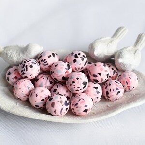 May include: A collection of pink terrazzo beads with black, gray, and red flecks, displayed on a white leaf-shaped dish. Two white bird figurines are positioned on the dish. The beads are round and smooth, perfect for jewelry making.