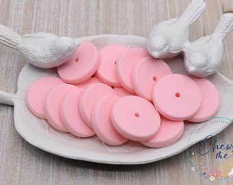 May include: A white ceramic dish shaped like a leaf holds a collection of pink silicone teething rings. The rings are round with a small hole in the center. The dish is on a light wood surface.