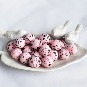 May include: A collection of pink terrazzo beads with black, gray, and red flecks, displayed on a white leaf-shaped dish. Two white bird figurines are positioned on the dish. The beads are round and appear to be approximately 0.75 inches in diameter.
