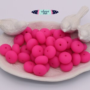 May include: A white ceramic dish with two white bird figurines on the edge, filled with bright pink silicone beads. The beads are round and have a hole in the center.