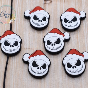 May include: Seven black, white, and red Jack Skellington-themed charms, each wearing a Santa hat. The charms are arranged on a light brown wooden surface. The charms are likely for shoes or other accessories.