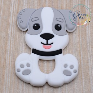 May include: A white and gray silicone teether shaped like a dog with black eyes, a black nose, and a pink tongue. The teether has a ring in the middle for easy gripping.