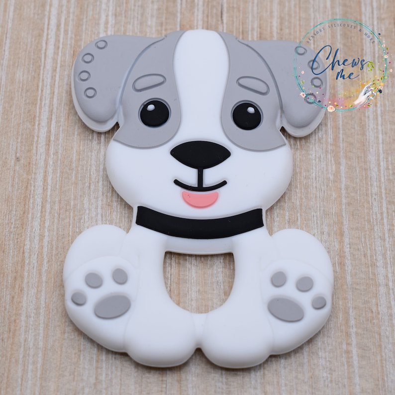 May include: A white and gray silicone teether shaped like a dog with black eyes, a black nose, and a pink tongue. The teether has a ring in the middle for easy gripping.