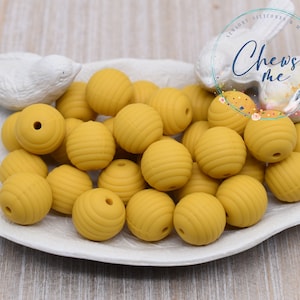 May include: A white ceramic dish with a leaf design holds a collection of 20 yellow silicone beads. The beads have a textured, ribbed surface. The dish is on a wooden surface.