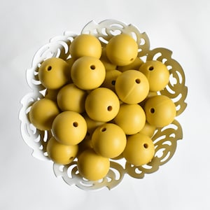 May include: A white bowl filled with mustard yellow silicone beads. The bowl has a decorative, ornate design.