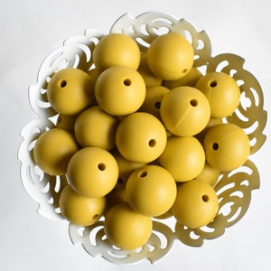 May include: A bowl filled with mustard yellow silicone beads. The beads are smooth and round, and they are arranged in a random pattern.