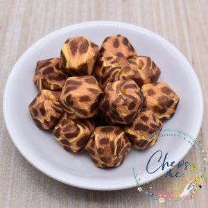 May include: A close-up of a pile of brown and yellow silicone chew beads with a woodgrain pattern. The beads are shaped like irregular hexagons with a hole in the center. The beads are on a white plate.