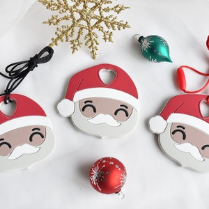 May include: Three red and white silicone Santa Claus ornaments with a heart-shaped cutout on the hat. Each ornament has a black cord attached to it. Two of the ornaments are on a white surface, and the third ornament is hanging from a red cord.