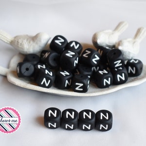 May include: A pile of black silicone baby teethers shaped like cubes with the letter "Z" printed in white on each side. The teethers are arranged on a white ceramic dish shaped like a bird.