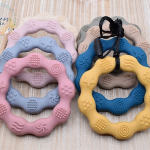 May include: A set of six colorful silicone teething rings with different textured surfaces. Each ring has a variety of shapes and patterns, including stars, waves, and circles. The rings are connected by a black cord.