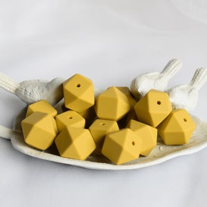 May include: A group of mustard yellow silicone teething beads in a geometric shape with a hole in the center. The beads are arranged in a white ceramic dish.