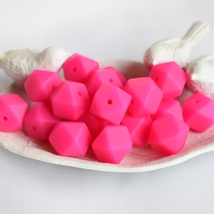 May include: A close-up of a pile of bright pink silicone beads. The beads are shaped like a hexagon with a hole in the center. The beads are on a white plate with two white bird figurines.