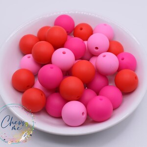 May include: A collection of colorful silicone beads in a white bowl. The beads are in shades of red, pink, and fuchsia. Each bead is round with a small hole. The bowl is white, and the background is a light color.