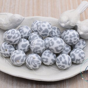 May include: A collection of gray and white silicone beads with a leopard print pattern. The beads are arranged in a white dish with two white bird figurines.