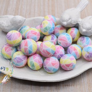 May include: A collection of colorful, pastel-toned silicone beads with a geometric pattern. The beads are arranged in a white ceramic dish shaped like a leaf. The dish is decorated with a small bird figurine.