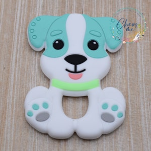 May include: A white silicone teether shaped like a dog with teal ears, a green collar, and gray paw prints. The dog has black eyes and a pink nose.