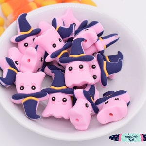 May include: A white bowl filled with pink ghost-shaped beads wearing navy blue witch hats with yellow accents. The beads have black eyes and smiling faces. Candy corn is visible in the background. The logo "chews me" is in the bottom right corner.