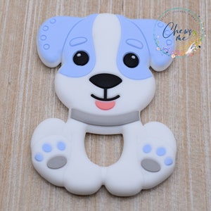 May include: A white silicone teether shaped like a dog with blue ears and a gray nose. The teether has a ring in the middle for easy gripping.