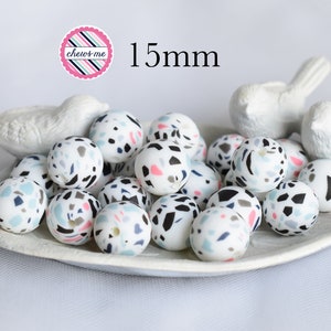 May include: A close-up of a white bowl filled with 15mm silicone beads. The beads are white with a terrazzo pattern in black, pink, and blue. The text "chew-me" is visible in the top left corner of the image. The text "15mm" is visible in the top center of the image.