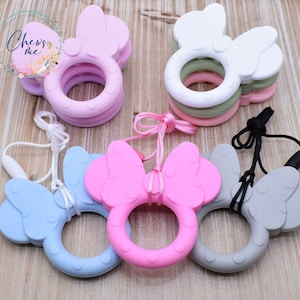 K&ouml;nnte beinhalten: Vier Silikon-Bei&szlig;ringe in Form einer Schleife mit Ohren. Die Bei&szlig;ringe sind in Pink, Blau, Grau und Wei&szlig;. Jeder Bei&szlig;ring hat eine passende Schnur.