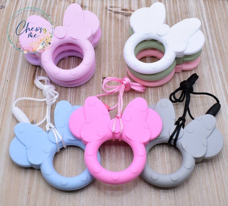 K&ouml;nnte beinhalten: Vier Silikon-Bei&szlig;ringe in Form einer Schleife mit Ohren. Die Bei&szlig;ringe sind in Pink, Blau, Grau und Wei&szlig;. Jeder Bei&szlig;ring hat eine passende Schnur.