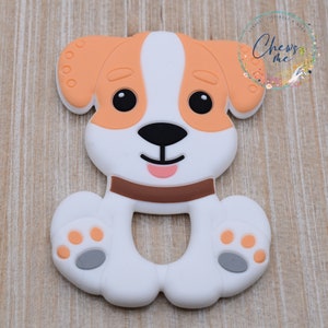 May include: A white and orange silicone teether shaped like a dog with brown eyes, a brown nose, and a pink tongue. The dog has a brown collar and gray paw prints on its feet. "Chews Me" is written in a circle around the dog.
