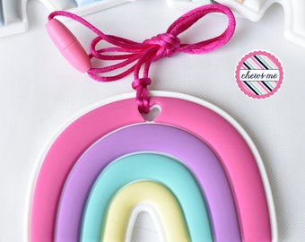 May include: A colorful rainbow-shaped teething toy with a pink cord. The rainbow features layers of pink, purple, blue, and yellow. The toy has a white border and a heart-shaped hole at the top. The text "chews me" is on a pink and blue sticker.