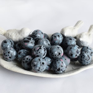 May include: A pile of gray and blue patterned beads in a white ceramic dish shaped like a leaf. The beads are round and have a textured surface.