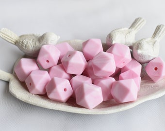 May include: A collection of pink silicone baby teethers in a geometric shape. The teethers are arranged in a white dish with two white bird figurines.