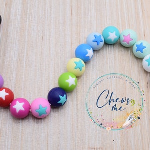Puede incluir: Una cadena colorida de cuentas de silicona con estrellas blancas. Las cuentas est&aacute;n en un patr&oacute;n de arco&iacute;ris, con cada cuenta de un color diferente. Las cuentas est&aacute;n enhebradas en una cuerda blanca. El texto "Chews Me" es visible en la imagen.