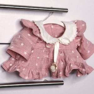 Puede incluir: Un vestido de muñeca rosa con patrones de estrellas blancas y un cuello blanco con ribete de encaje. El vestido tiene mangas con volantes y una cinta con un dije de perla y oro. Está colgado de una percha blanca.