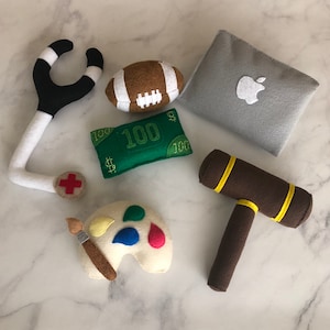 May include: A collection of felt toys representing various professions. Includes a stethoscope, football, laptop with an apple logo, a felt $100 bill, a gavel, and a painter's palette with a brush.