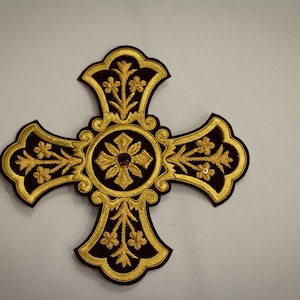 May include: A black velvet cross with gold embroidery, featuring a central floral design with a red jewel in the center.