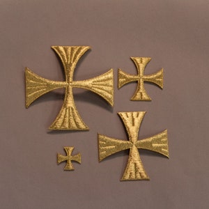 May include: A collection of gold-colored embroidered crosses in various sizes. The crosses have a detailed, textured design and are arranged on a neutral gray background. The embroidery work is visible, highlighting the craftsmanship.