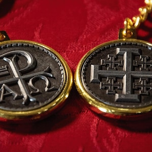 Puede incluir: Dos colgantes circulares con borde dorado y diseños en negro y plata. Un colgante presenta una cruz con cruces más pequeñas en cada extremo. El otro colgante presenta una letra P estilizada con una corona encima y las letras A y O a cada lado.