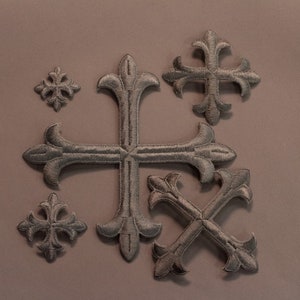 May include: Six silver-colored, ornate, cross-shaped embellishments with a fleur-de-lis design. The crosses are arranged in a cluster on a light brown background.