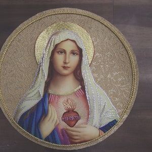 Puede incluir: Una imagen redonda con marco dorado de la Virgen María con un corazón en el pecho. El corazón es rojo y tiene una corona de espinas. La imagen está sobre un fondo de madera marrón.