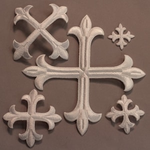 May include: A selection of white embroidered crosses in different sizes. Each cross has a fleur-de-lis design at the ends of the arms. The embroidery has a textured look, and the crosses are displayed on a neutral background.
