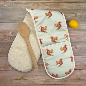 Könnte beinhalten: Ein Paar Ofenhandschuhe mit hellgrünem Hintergrund und einem Muster aus roten Eichhörnchen, die Nüsse halten. Ein Handschuh ist aus cremefarbenem Frottee.